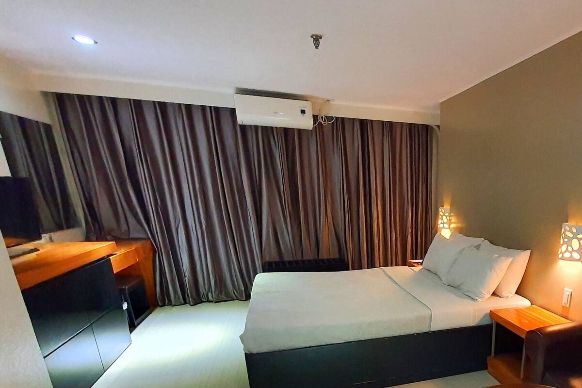 A hotel room with a bed, bedside lamps, a desk, a chair, and a painting on the wall.