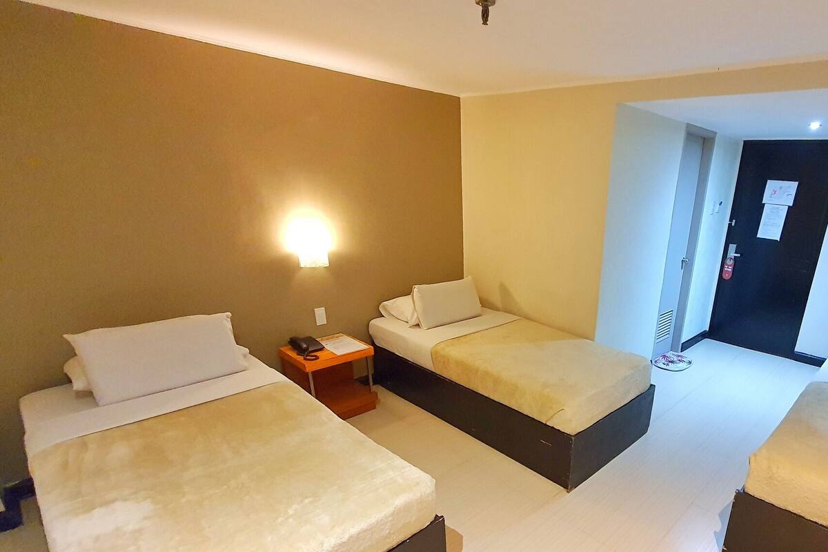 A hotel room with two beds, a nightstand, a wall-mounted light, and a door with a fire alarm.