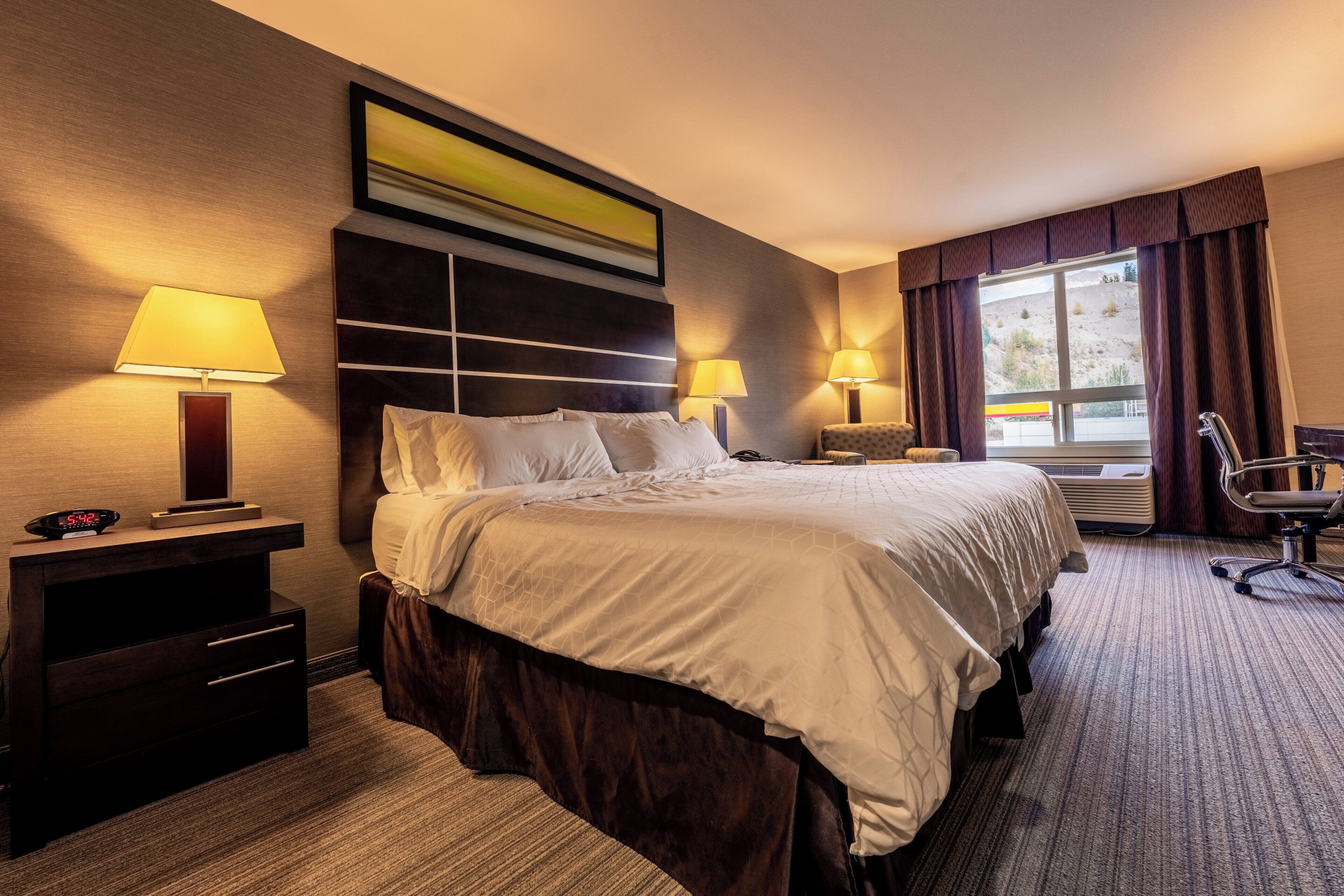 Quarto de hotel com uma cama grande, duas mesas de cabeceira, uma escrivaninha, uma cadeira e uma janela com cortinas.