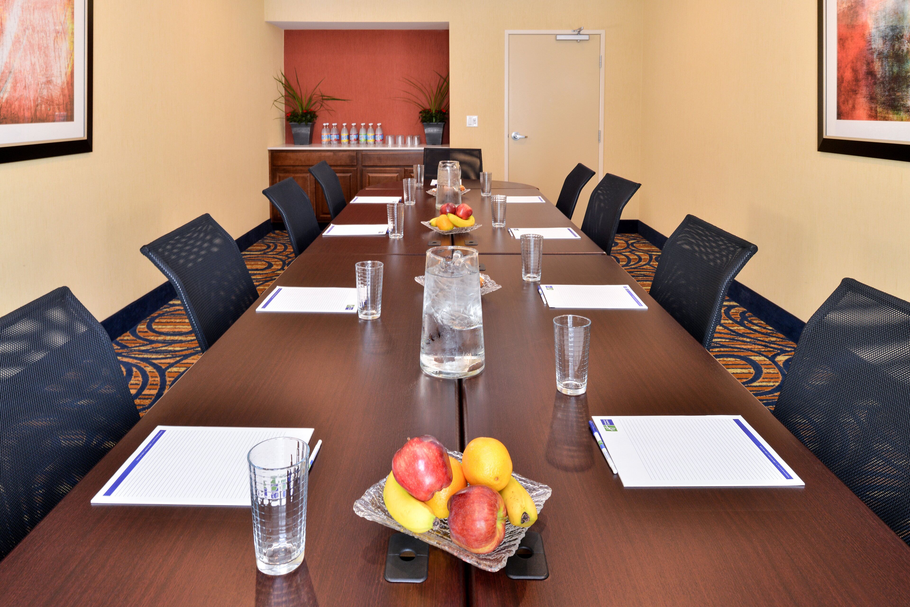 Un salón de conferencias con una mesa larga, sillas, vasos de agua y una cesta de frutas.