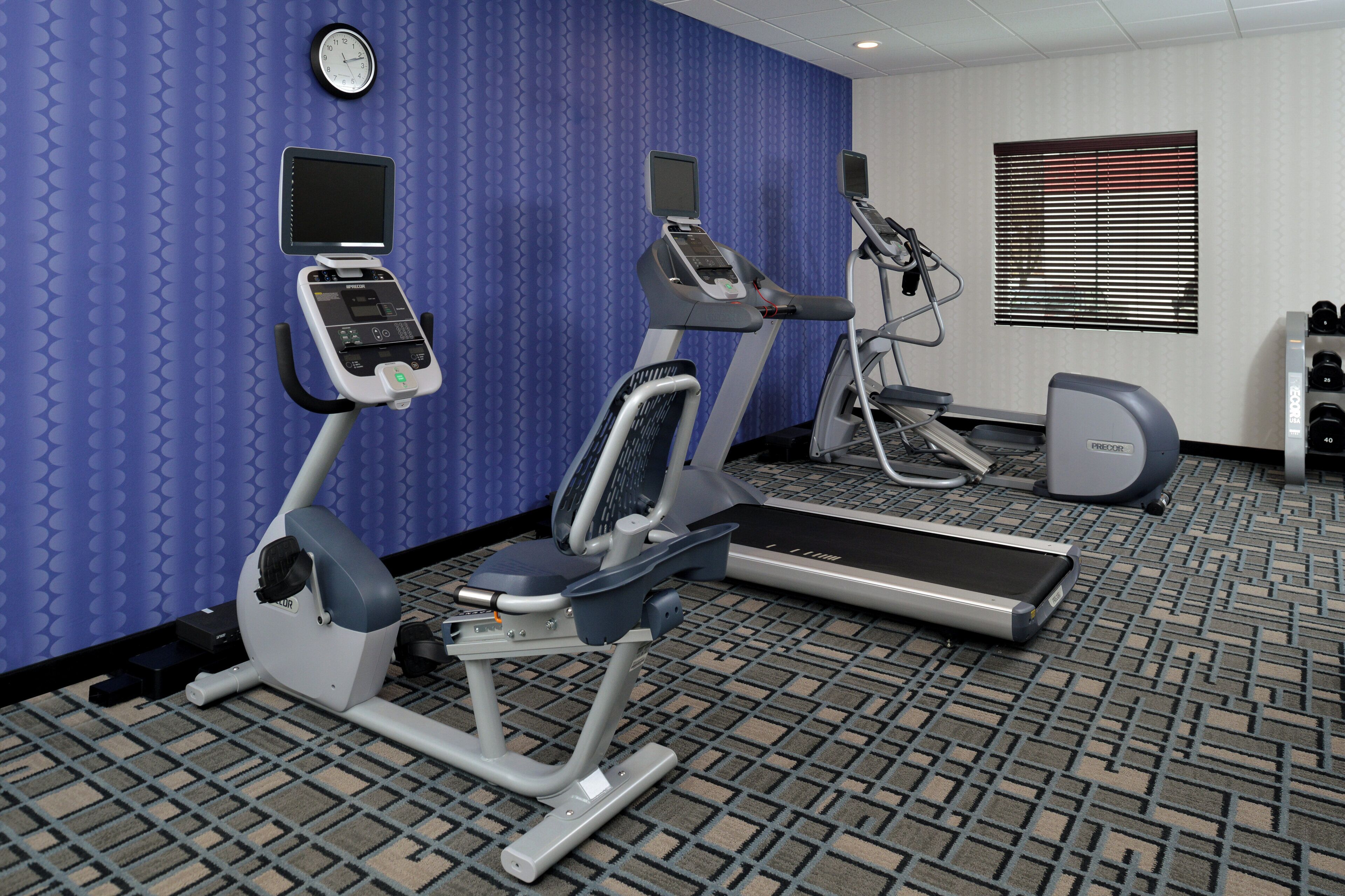 Un gimnasio con equipo de cardio, un reloj de pared y una ventana con persianas.