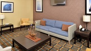A hotel room with a blue sofa, a glass coffee table, a patterned carpet, and framed artwork on the walls. - Holiday Inn Express & Suites Peekskill-Lower Hudson Valley by IHG (Peekskill)