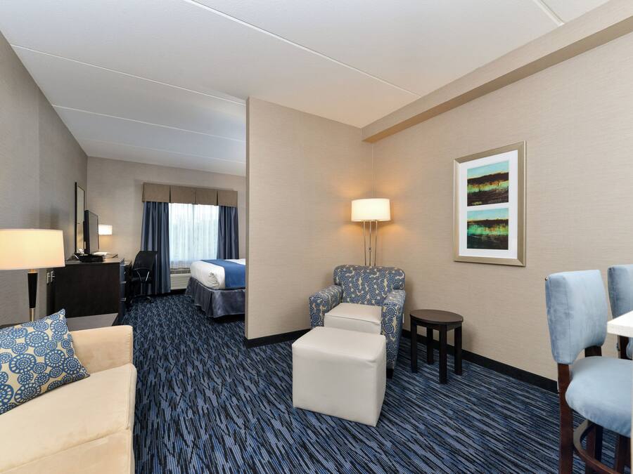 A hotel room with a bed, a sofa, a chair, a small table, a lamp, and a painting on the wall.