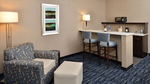 A hotel room with a patterned sofa, a small ottoman, a desk with two chairs, a microwave, a wine bottle, and a framed picture on the wall. - Holiday Inn Express & Suites Peekskill-Lower Hudson Valley by IHG (Peekskill)