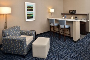 A hotel room with a patterned sofa, a small ottoman, a desk with two chairs, a microwave, a wine bottle, and a framed picture on the wall.