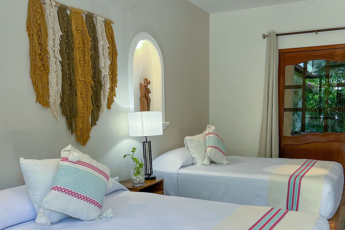 Habitación de hotel con dos camas, una mesita de noche con lámpara, un adorno en la pared y vistas a la vegetación a través de una ventana.