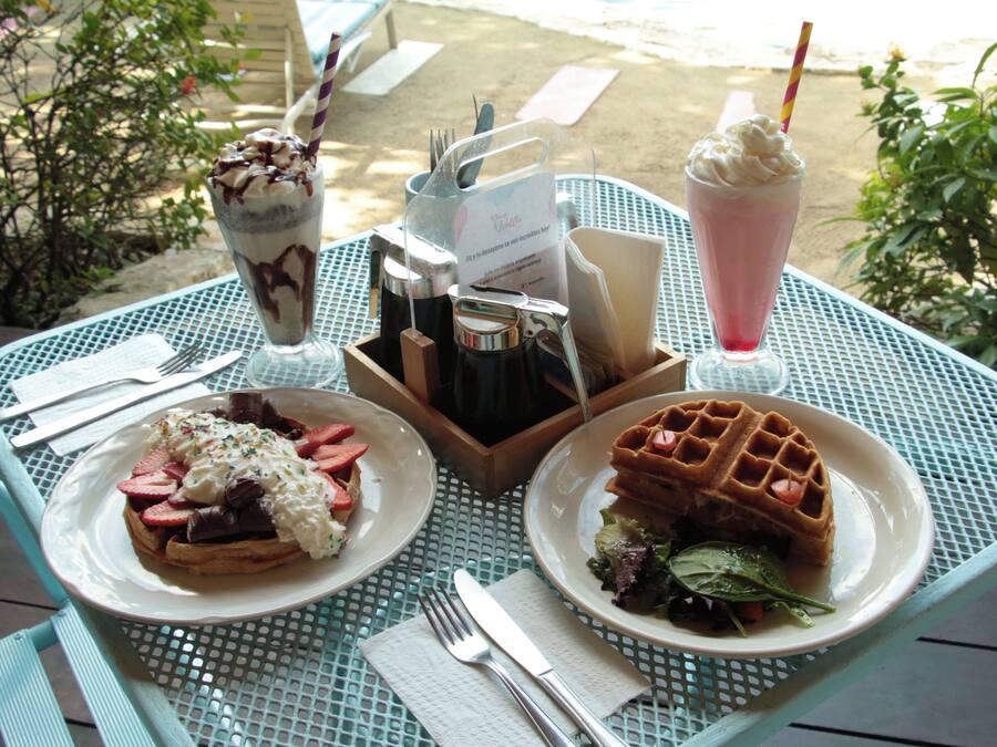 Una mesa dispuesta con un waffle, un plato de waffles con fresas, un postre con crema batida y chocolate, y una bebida rosada.