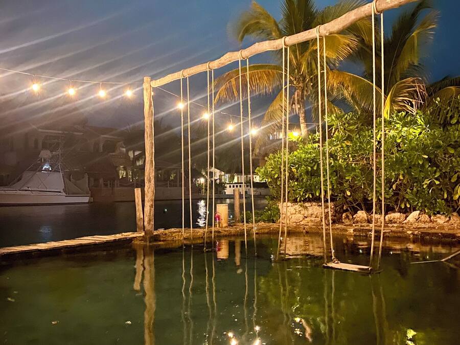 Vista nocturna de una piscina con luces colgantes, palmeras y un muelle.