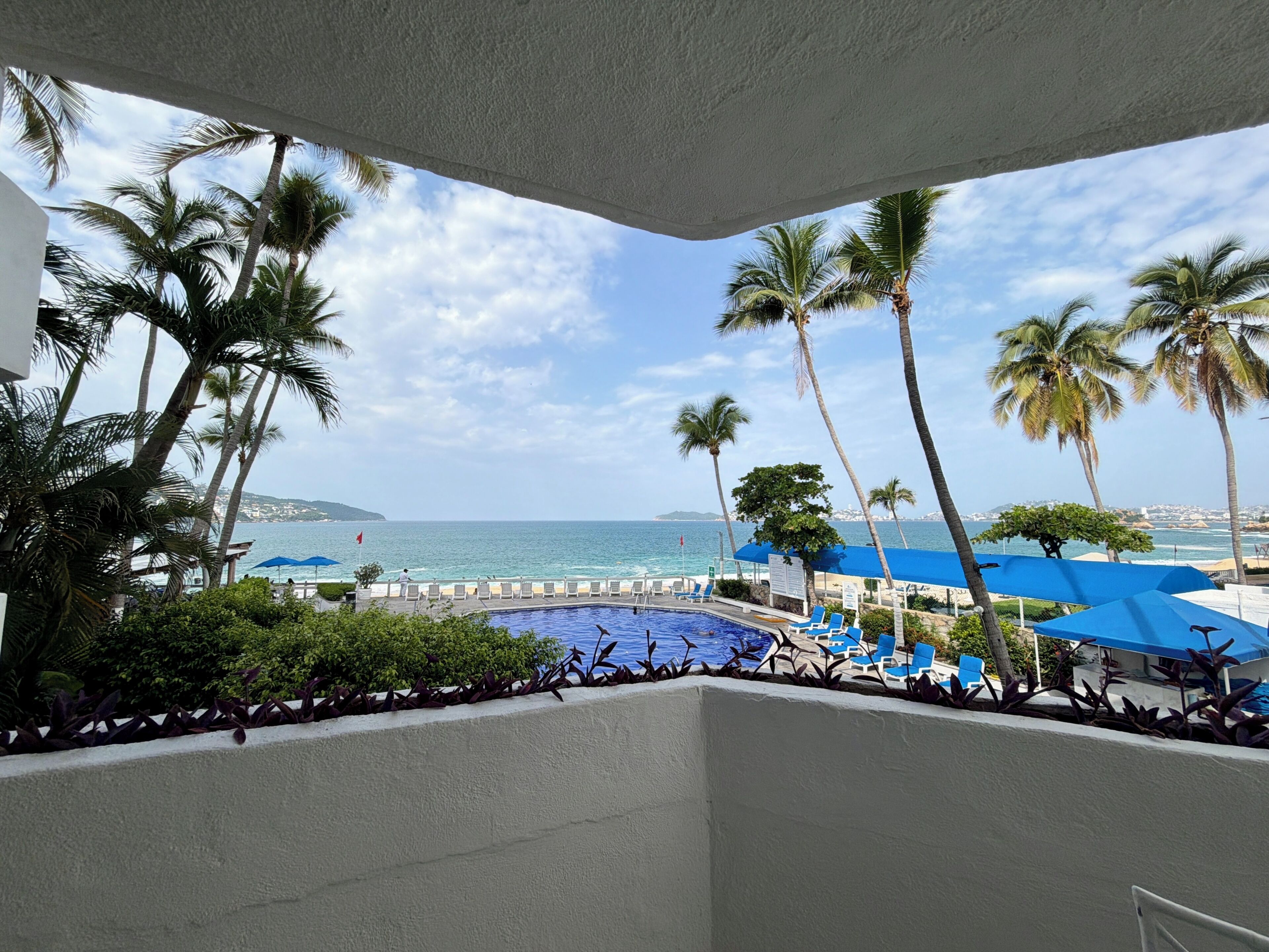 Zona de piscina con sillas y sombrillas azules, rodeada de palmeras y vistas al mar.