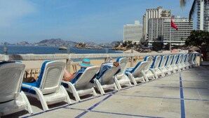 A row of beachside lounge chairs facing the ocean, with a clear view of the sea and distant buildings. - Hotel Acapulco Malibu (Acapulco)