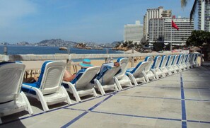 Una fila de sillas reclinables frente al mar, con vista despejada al océano y edificios distantes.