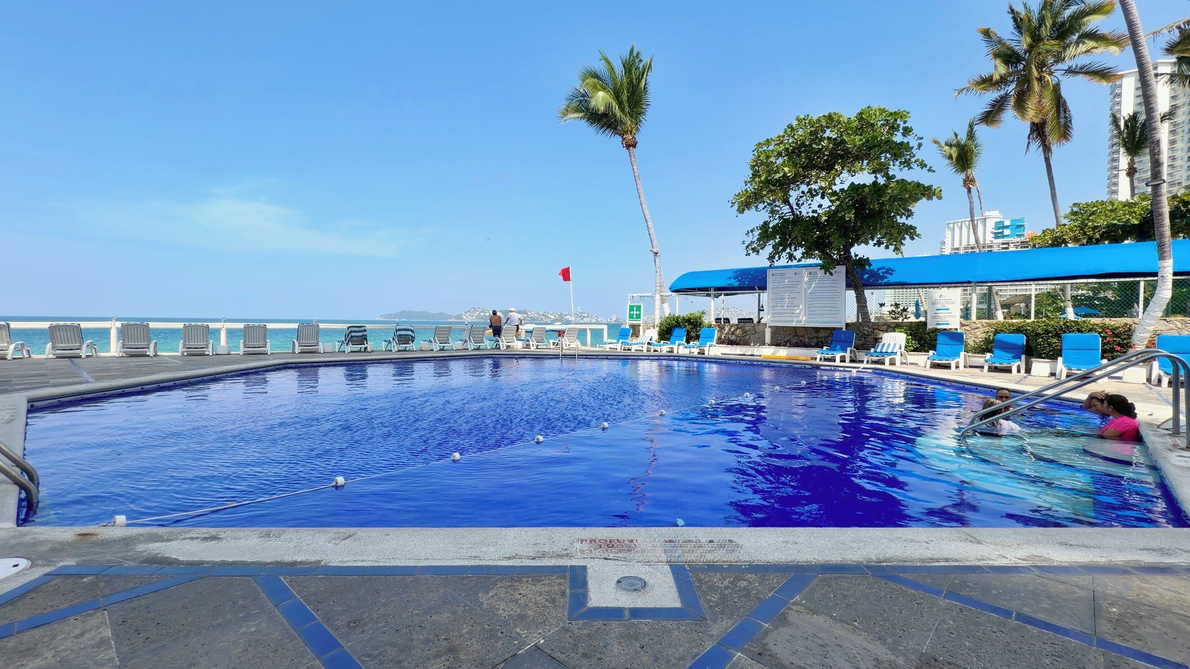 A swimming pool with lounge chairs and palm trees, a clear sky, and a distant view of the ocean.