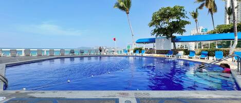A swimming pool with lounge chairs and palm trees, a clear sky, and a distant view of the ocean.