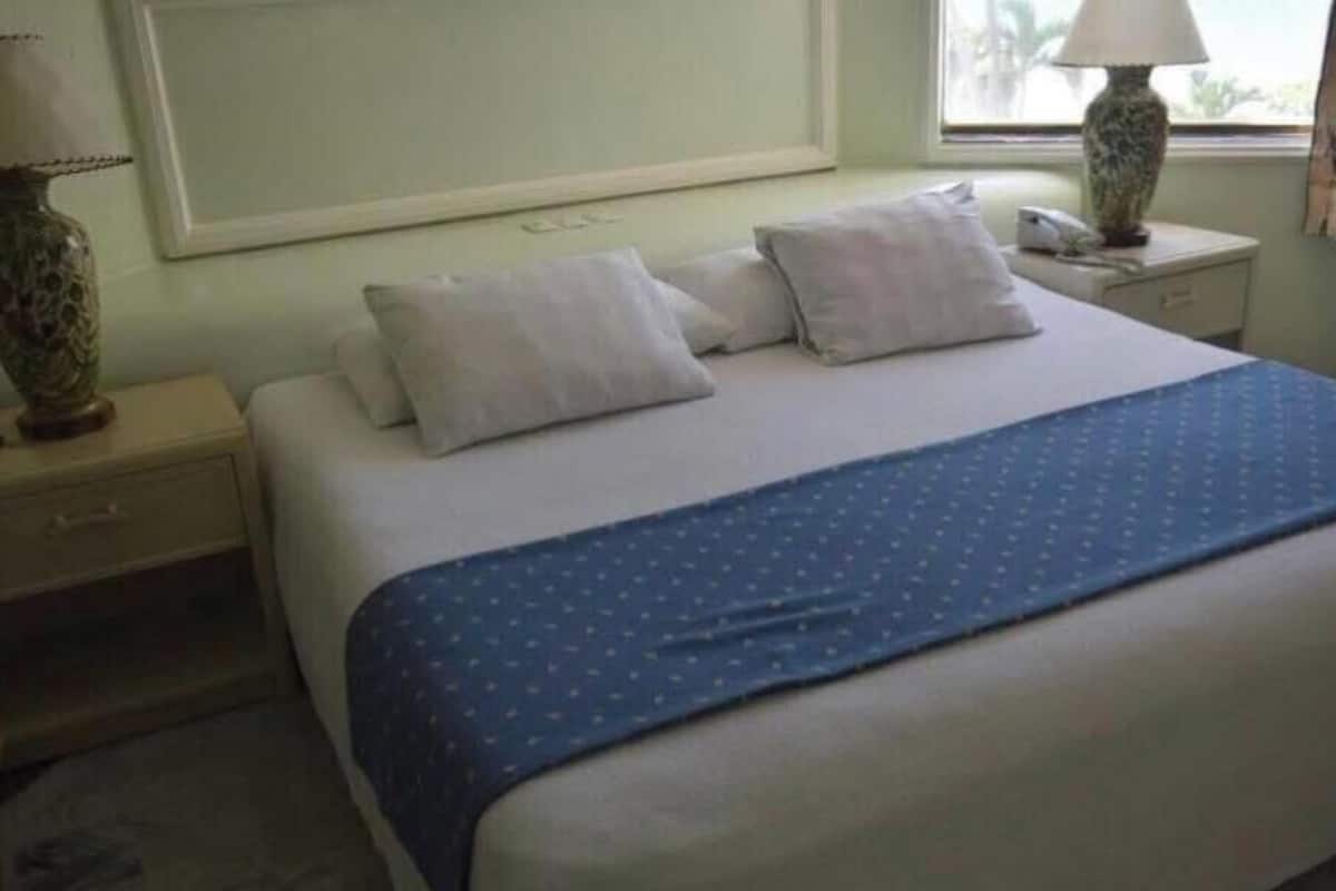 Una habitación de hotel con una cama, dos mesitas de noche, una lámpara y un ventanal con cortinas.