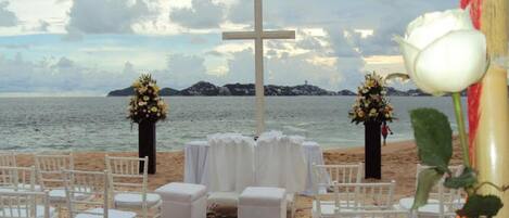 Une cérémonie de mariage organisée sur la plage, avec des chaises blanches et une croix au centre, le tout offrant une vue sur la mer.