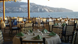 An outdoor dining area with tables set for two, overlooking a body of water with buildings in the background. - Hotel Acapulco Malibu (Acapulco)