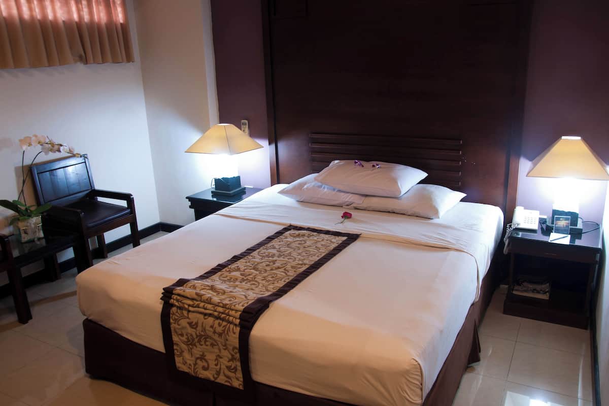 A neatly made bed with a decorative runner, two bedside lamps, a small table with a lamp, and a chair with a potted plant.