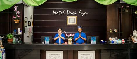 A reception desk at Hotel Puri Ayu with two staff members, a dark wall with the hotel name, and decorative elements like flowers and a framed certificate.