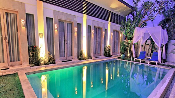 Una piscina rectangular amb superfície d'aigua blava cristal·lina, envoltada per un jardí ben cuidat i una filera de portes amb cortines.