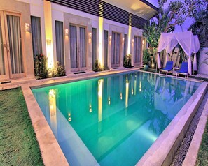 A rectangular swimming pool with a clear blue water surface, surrounded by a well-maintained lawn and a row of doors with curtains. - La Cabana Hotel and Villas (Keroboken Kelod)