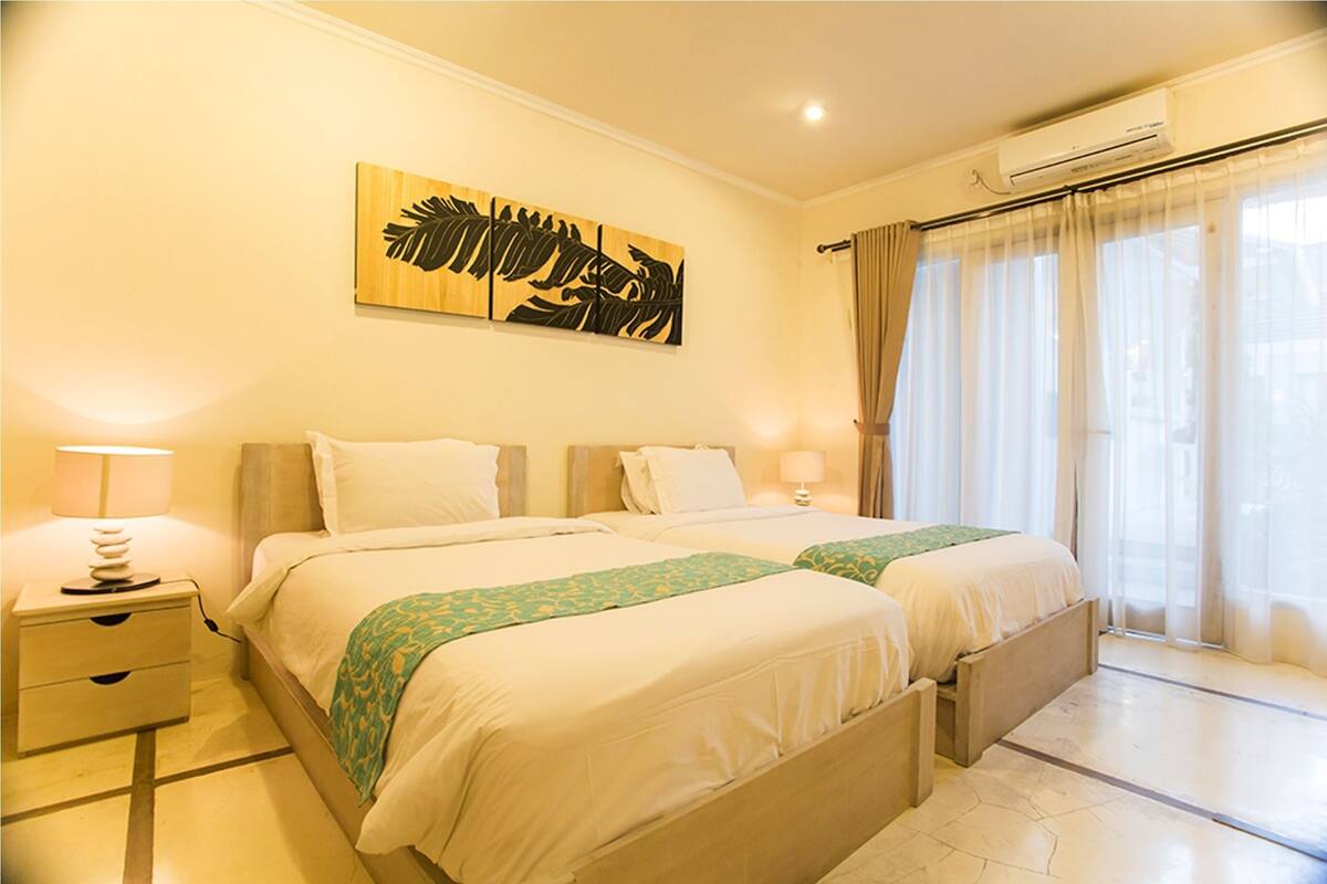 A hotel room with two beds, a nightstand with a lamp, a wall art piece, and a window with curtains.