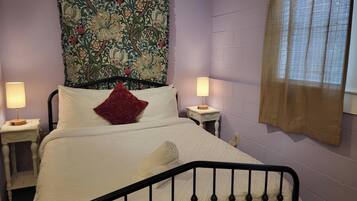 Une chambre à coucher avec un lit, deux tables de chevet, un oreiller rouge, une tapisserie et une fenêtre avec des rideaux.