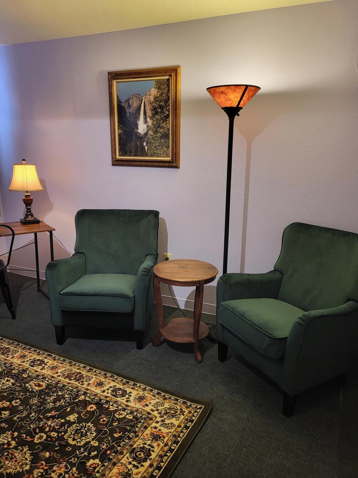 Una habitación con dos sillones verdes, una pequeña mesa redonda de madera, una lámpara de pie, un cuadro enmarcado de una cascada y una alfombra estampada.