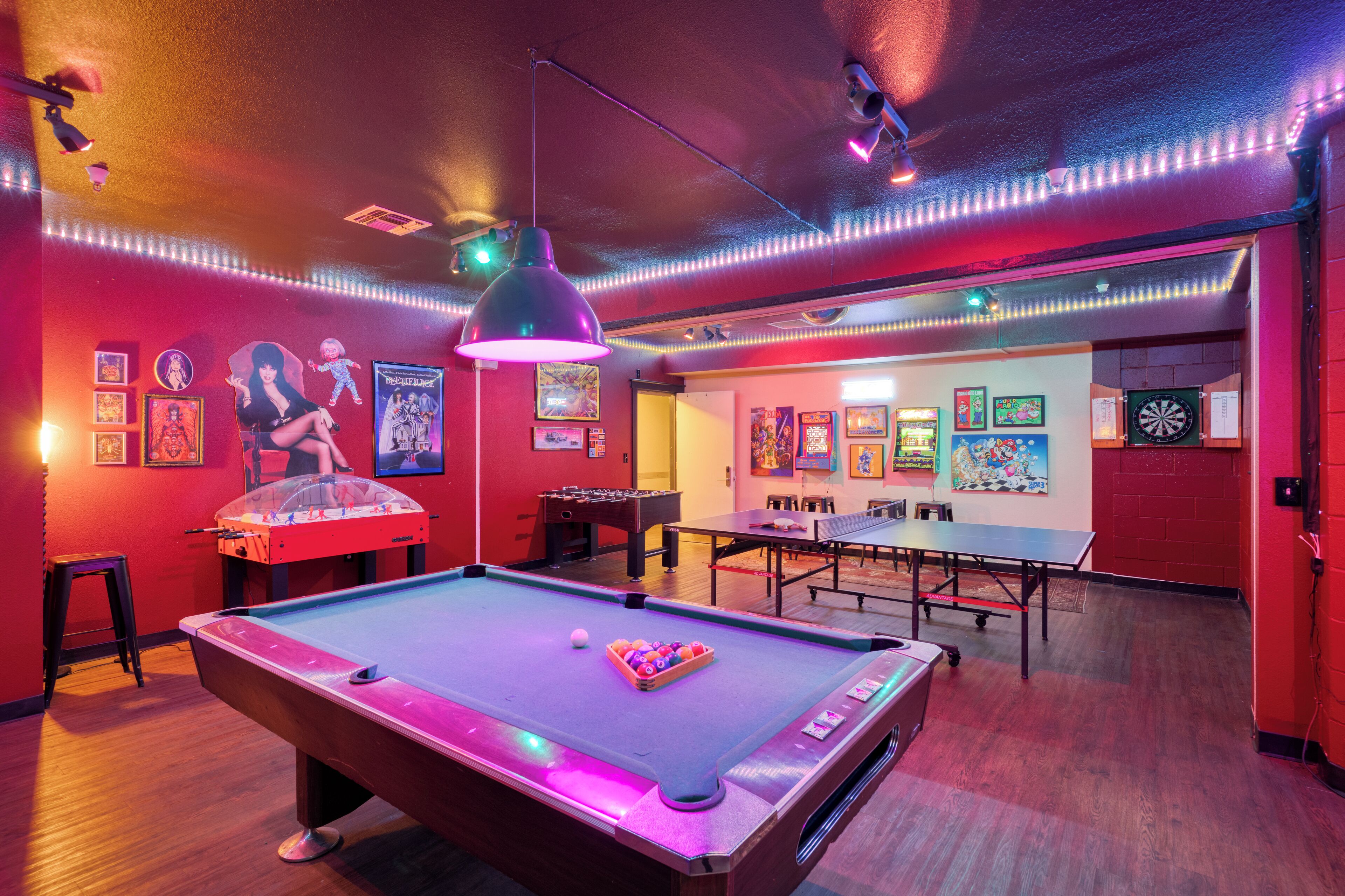 A pool table with a ball rack, a foosball table, and dartboards in a room with red walls and wooden floors.