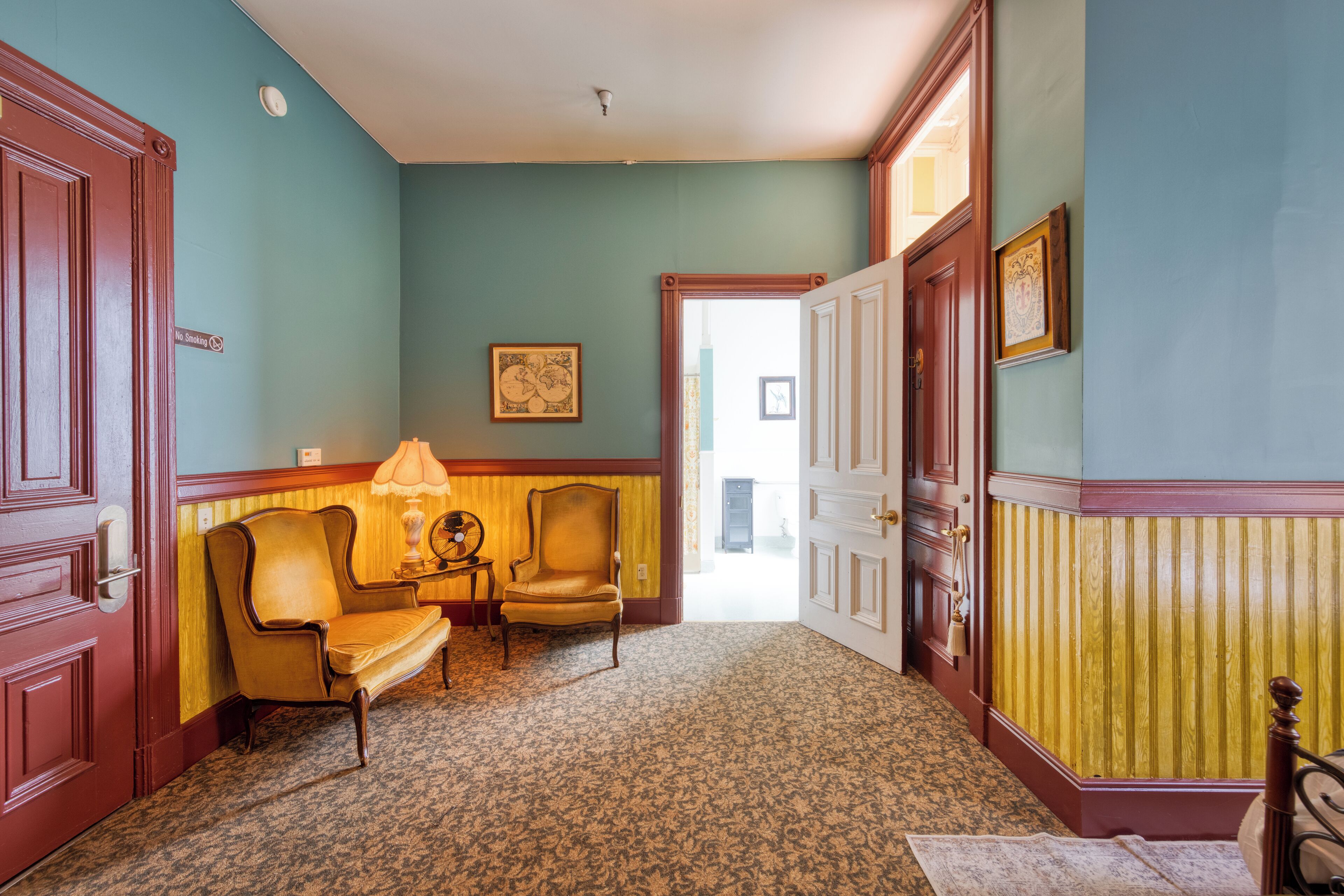 Un couloir avec un tapis à motifs, un canapé, deux fauteuils, une table avec une lampe et une porte sur laquelle est apposé un panneau « Interdiction de fumer ».