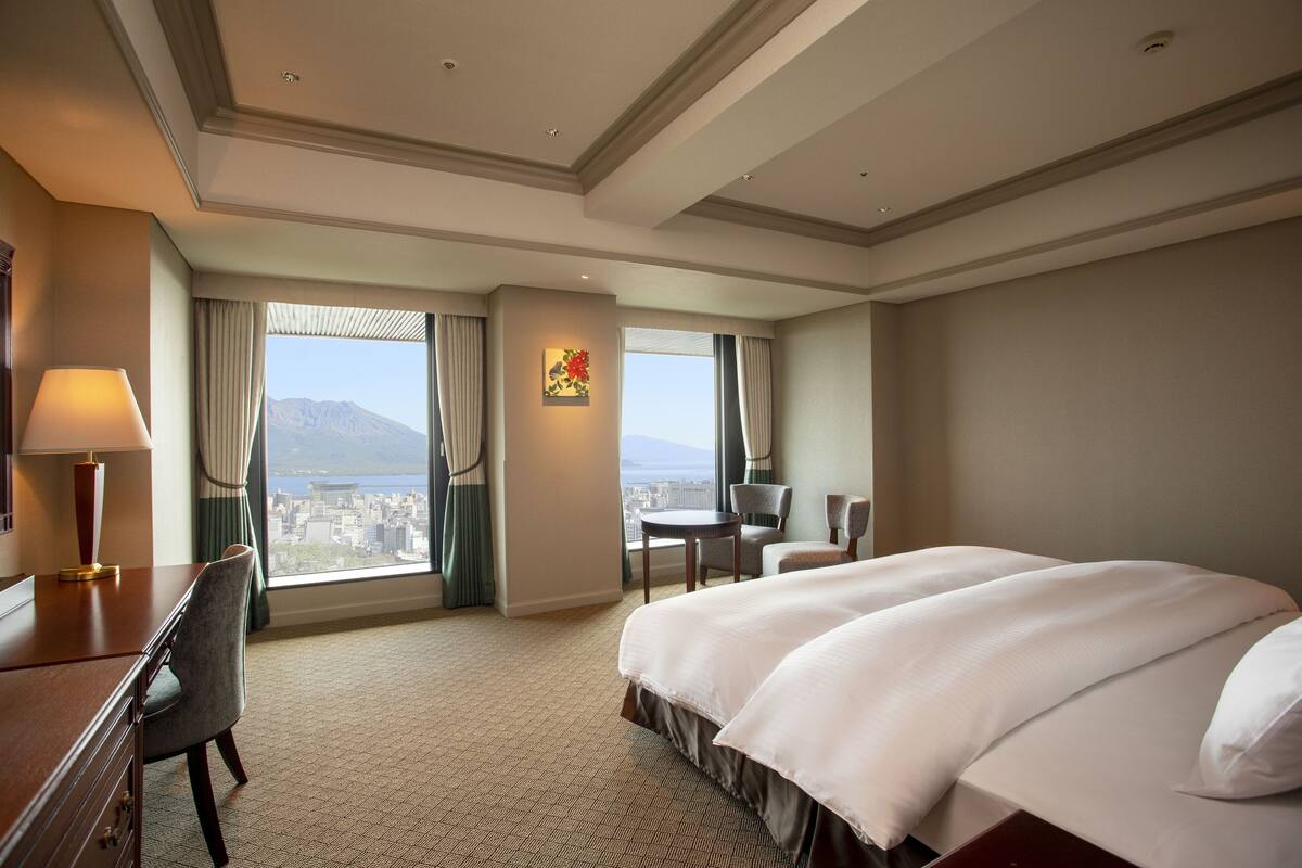A hotel room with a large bed, a desk, a chair, and a view of a cityscape through large windows.
