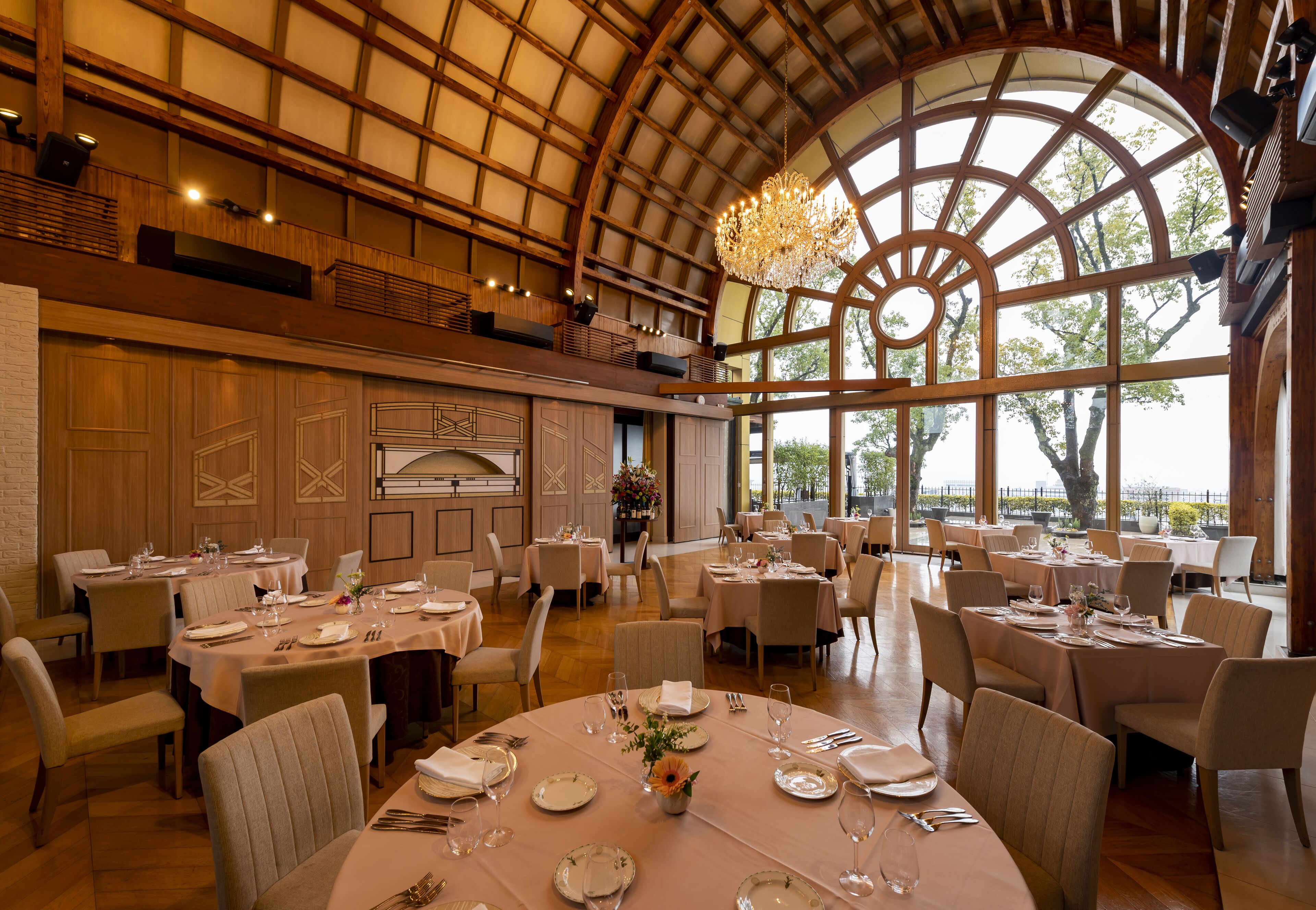 A spacious dining area with multiple tables set for guests, large windows offering natural light, and a high ceiling with wooden beams.