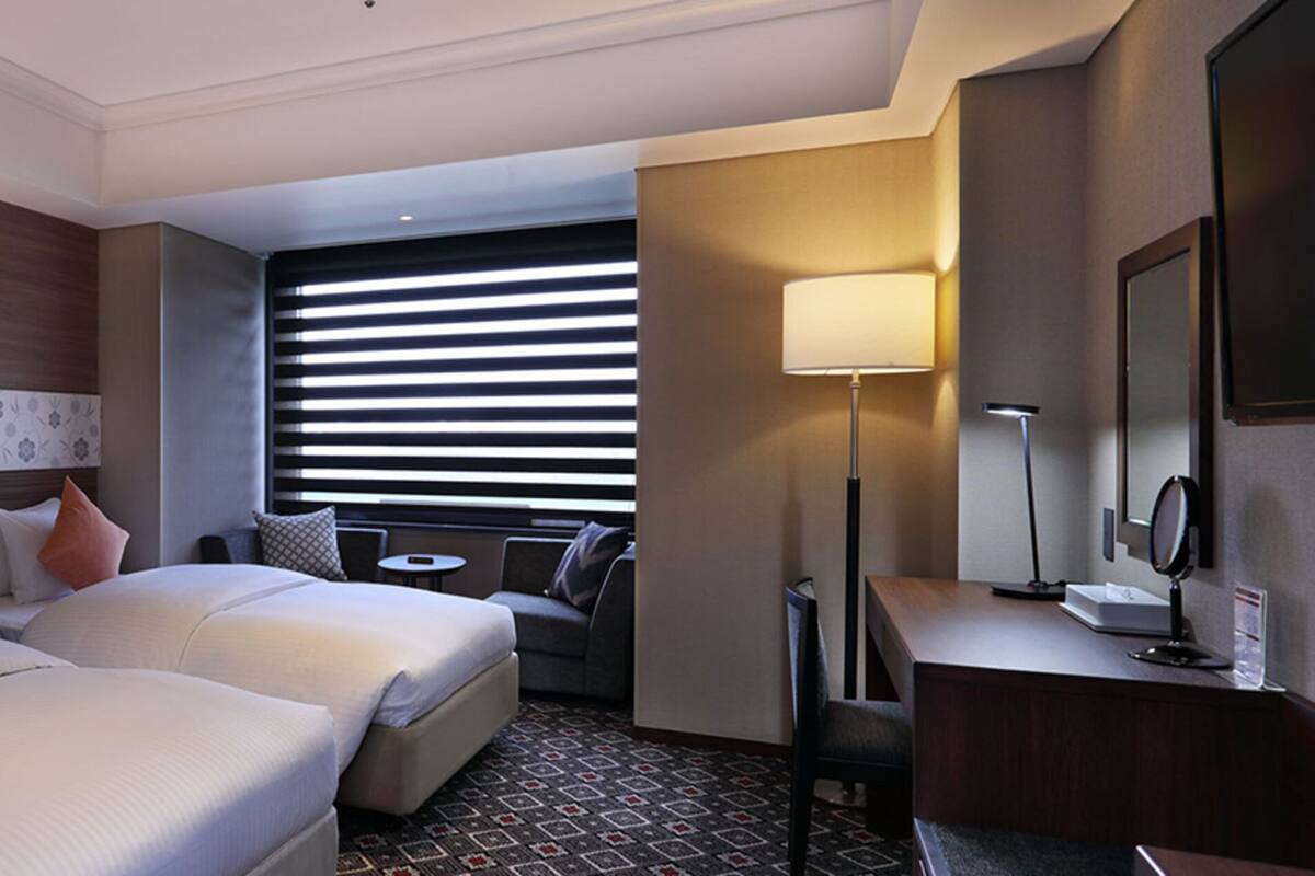 A hotel room with two beds, a desk, a chair, a lamp, and a window with blinds.