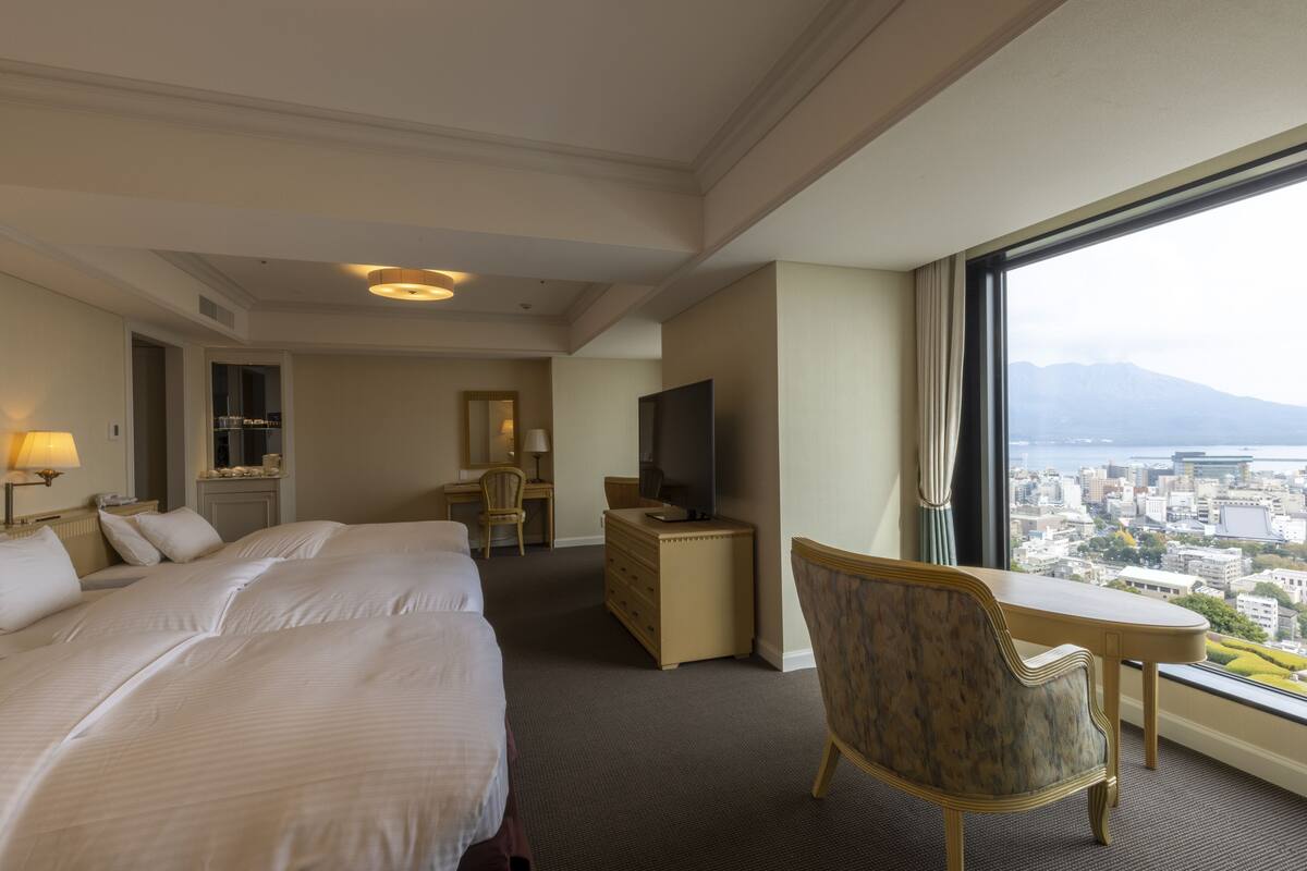 A hotel room with a large bed, a desk, a chair, a dresser, and a TV. There is a large window with a city view.