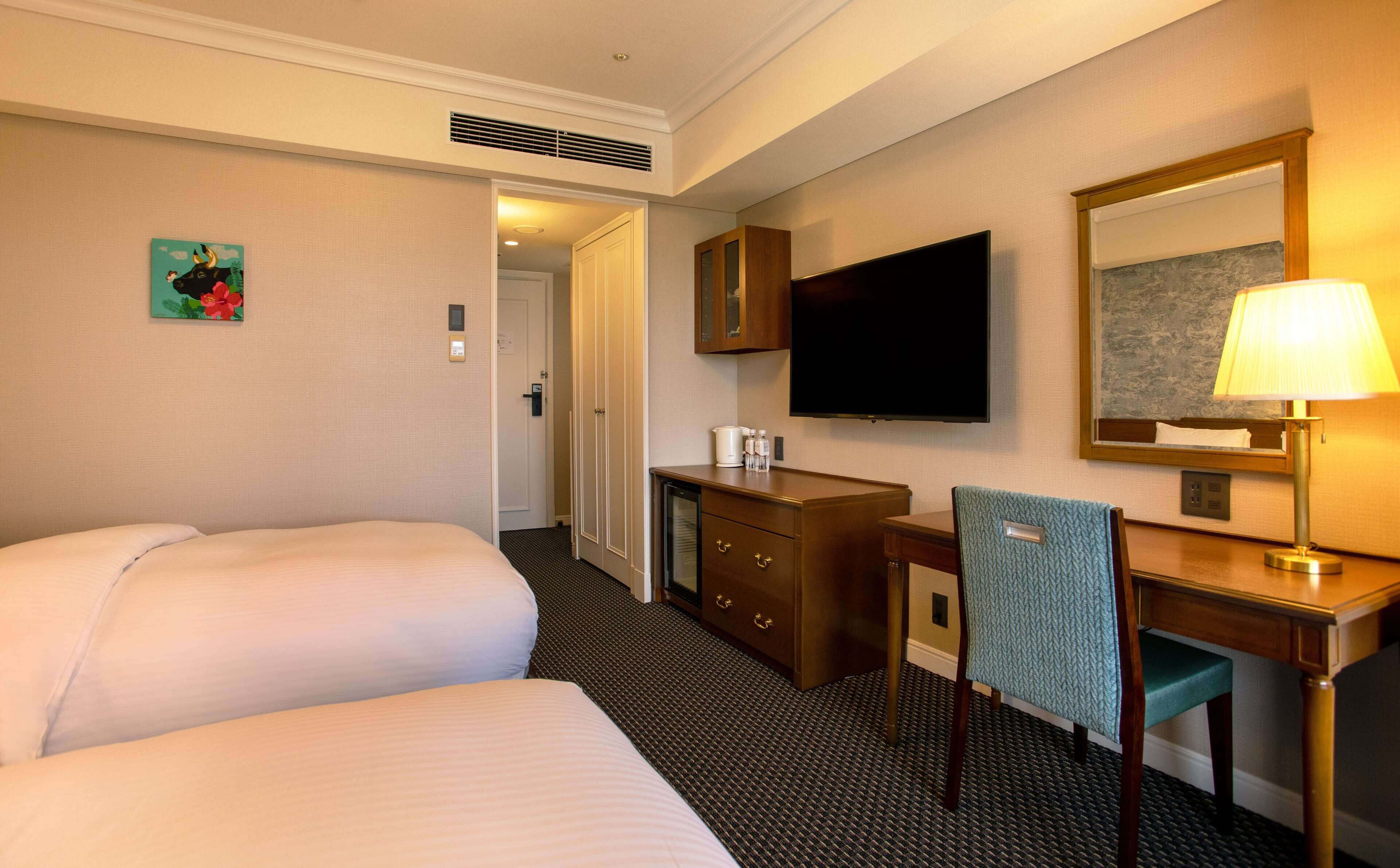 A hotel room with a bed, a desk with a chair, a television, a lamp, and a painting on the wall.