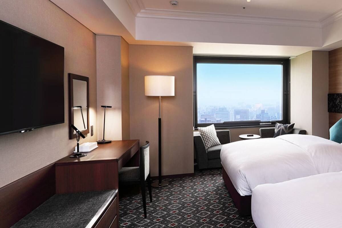 A hotel room with two beds, a desk, a chair, a TV, and a window with a city view.