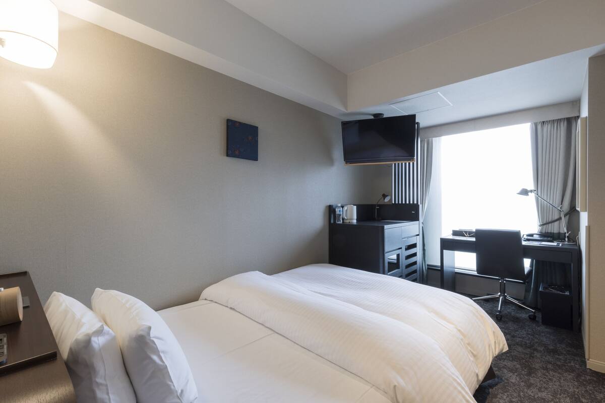 A hotel room with a large bed, a desk with a chair, a flat-screen TV mounted on the wall, and a window with curtains.