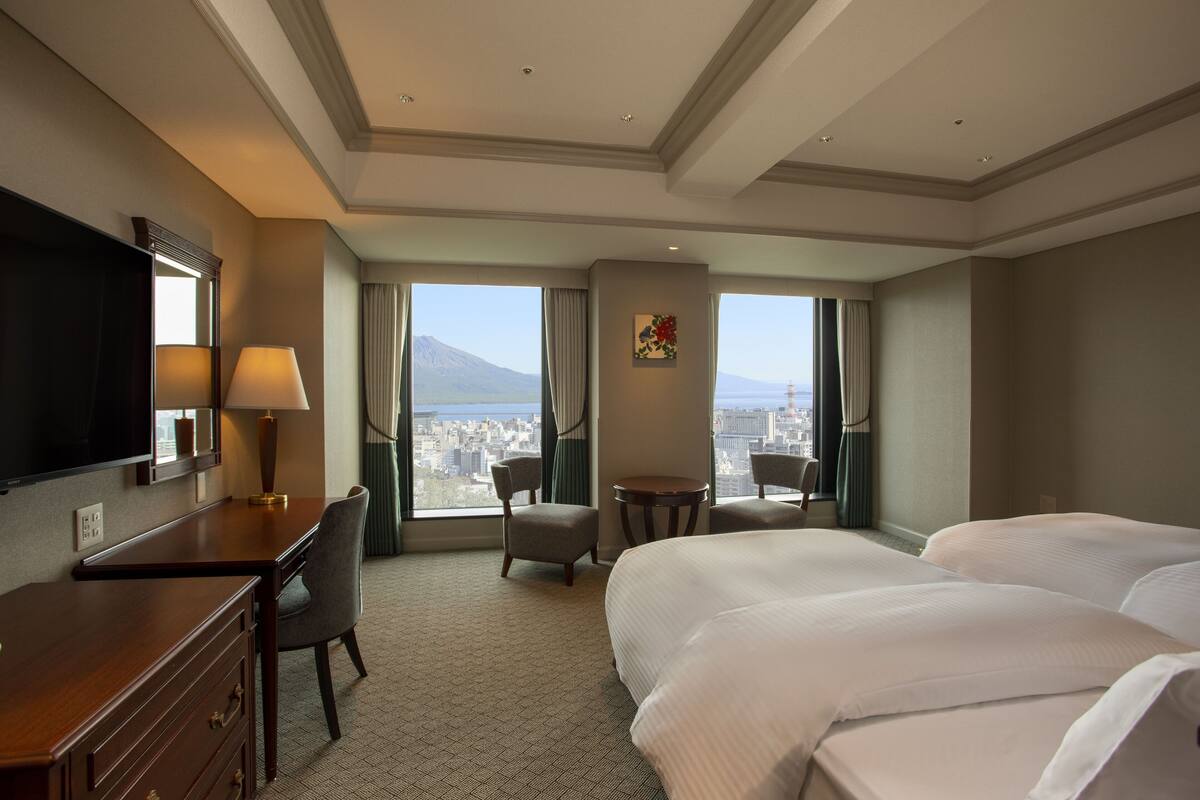 A hotel room with a large bed, a desk, a chair, a TV, and a view of the city and mountains.