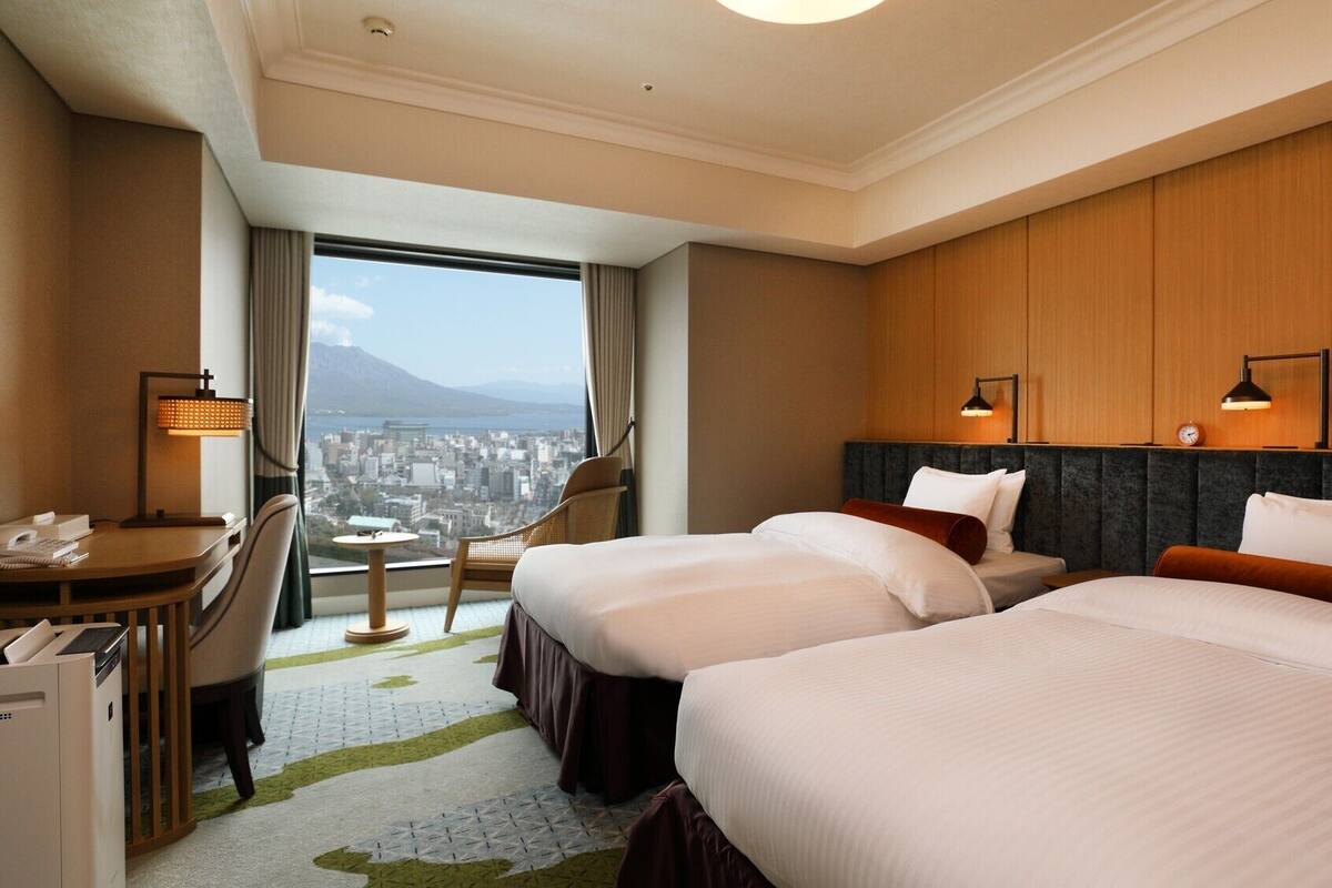 A hotel room with two beds, a desk, a chair, and a view of a cityscape.
