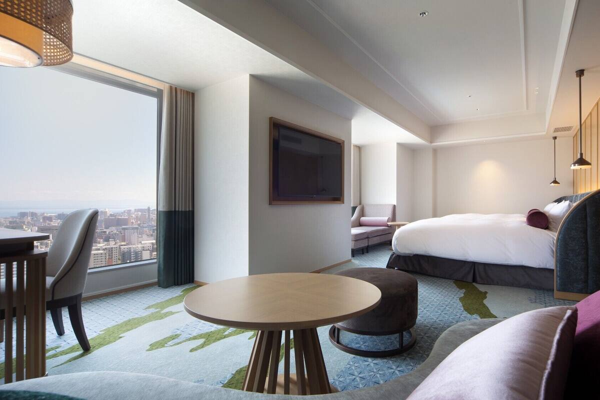 A hotel room with a large window, a bed, a sofa, a table, and a TV.