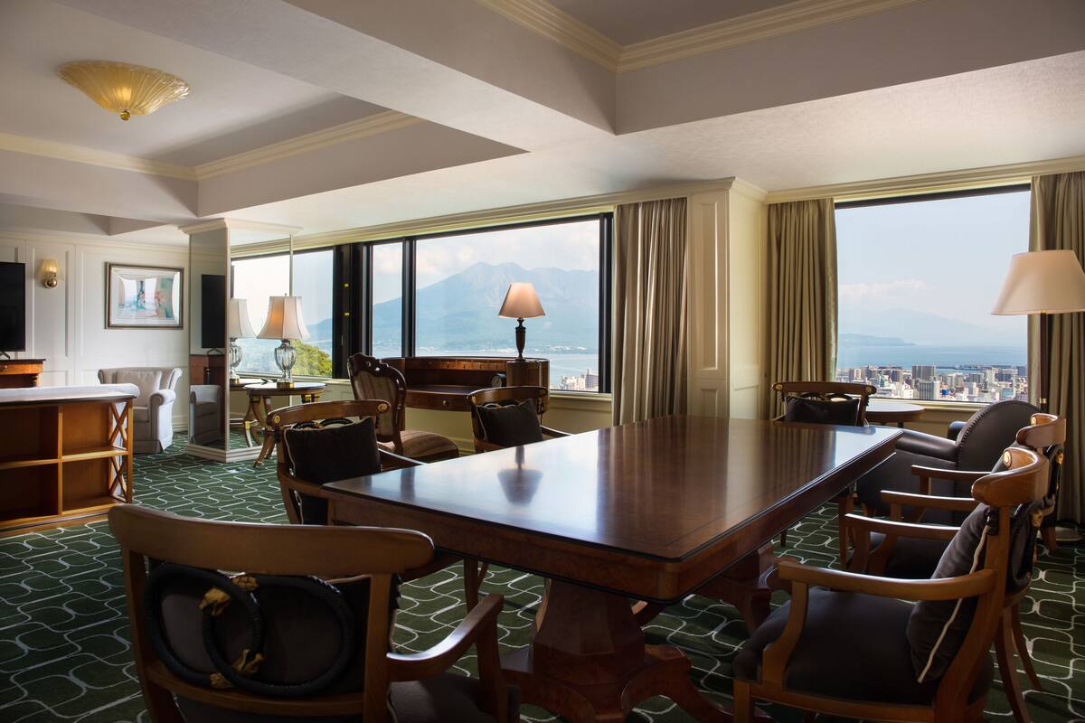 A hotel conference room with a large table, multiple chairs, a television, and a view of a cityscape through large windows.