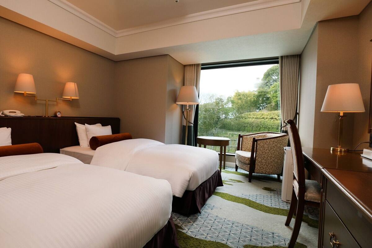 A hotel room with two beds, a desk, a chair, and a view of the outdoors.