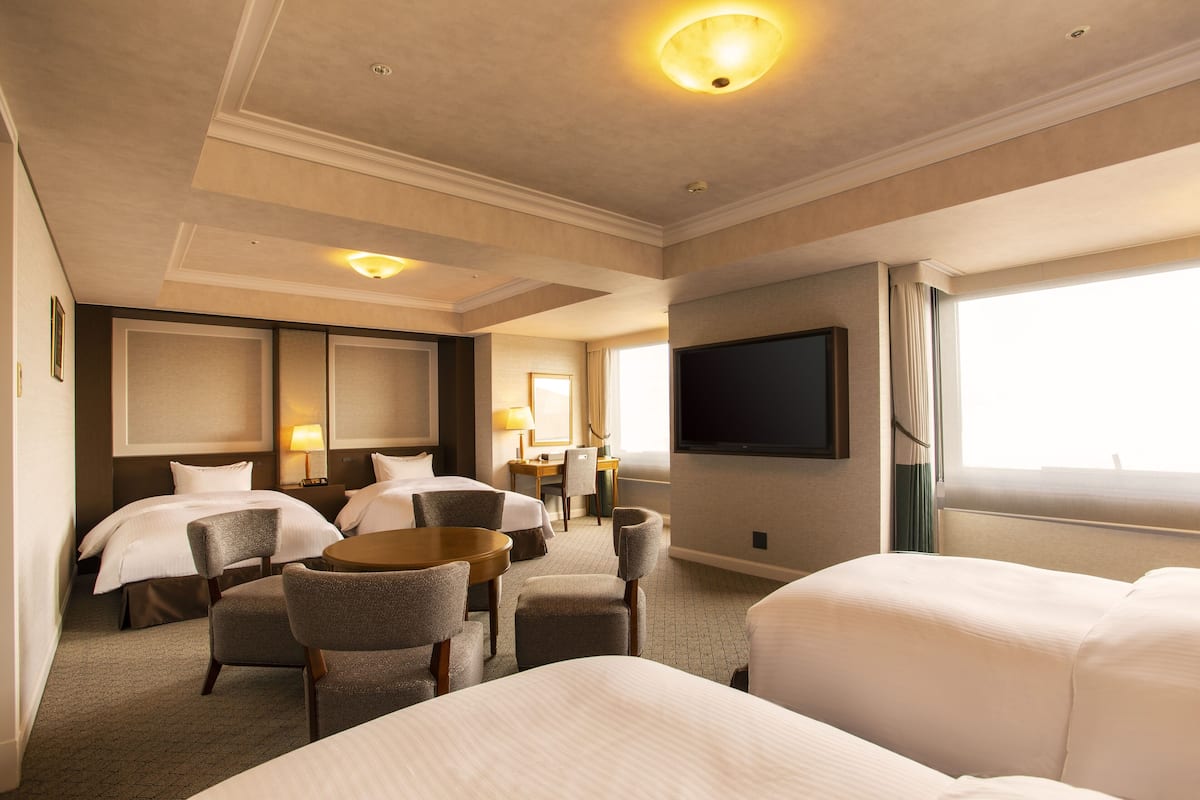 A hotel room with two beds, a desk, a chair, a flat-screen TV, and a large window.