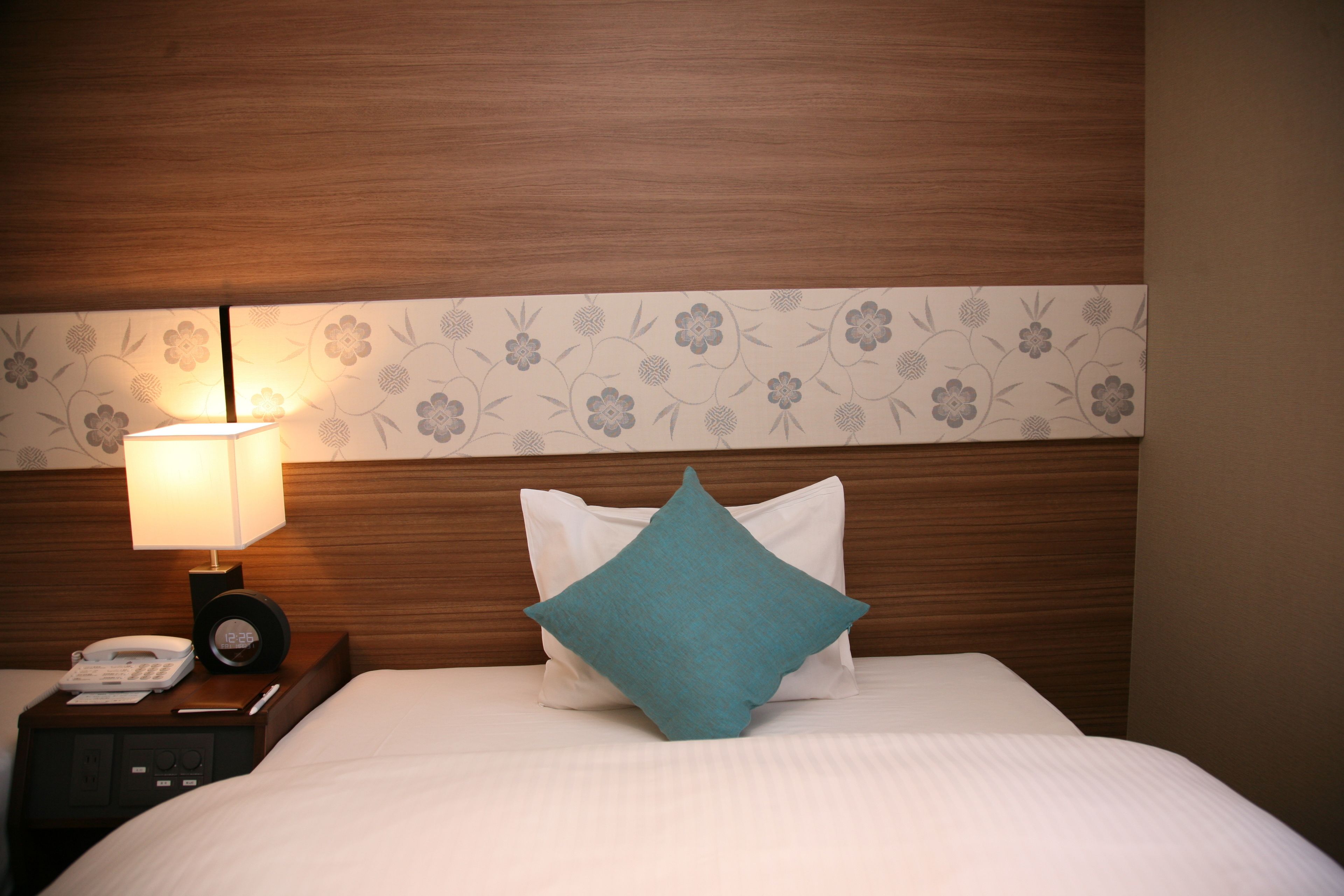 A neatly made bed with a teal pillow, a bedside table with a lamp, and a clock displaying 12:25.