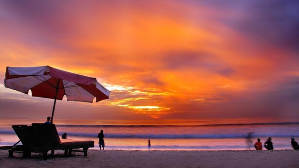A beach scene with a sunset, a beach umbrella, and people relaxing on the sand. - Kuta Sari House (Kuta)
