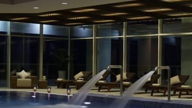 A modern indoor pool with multiple water features, glass walls, and lounge chairs. - Cocor Spa Hotel (Neptun)