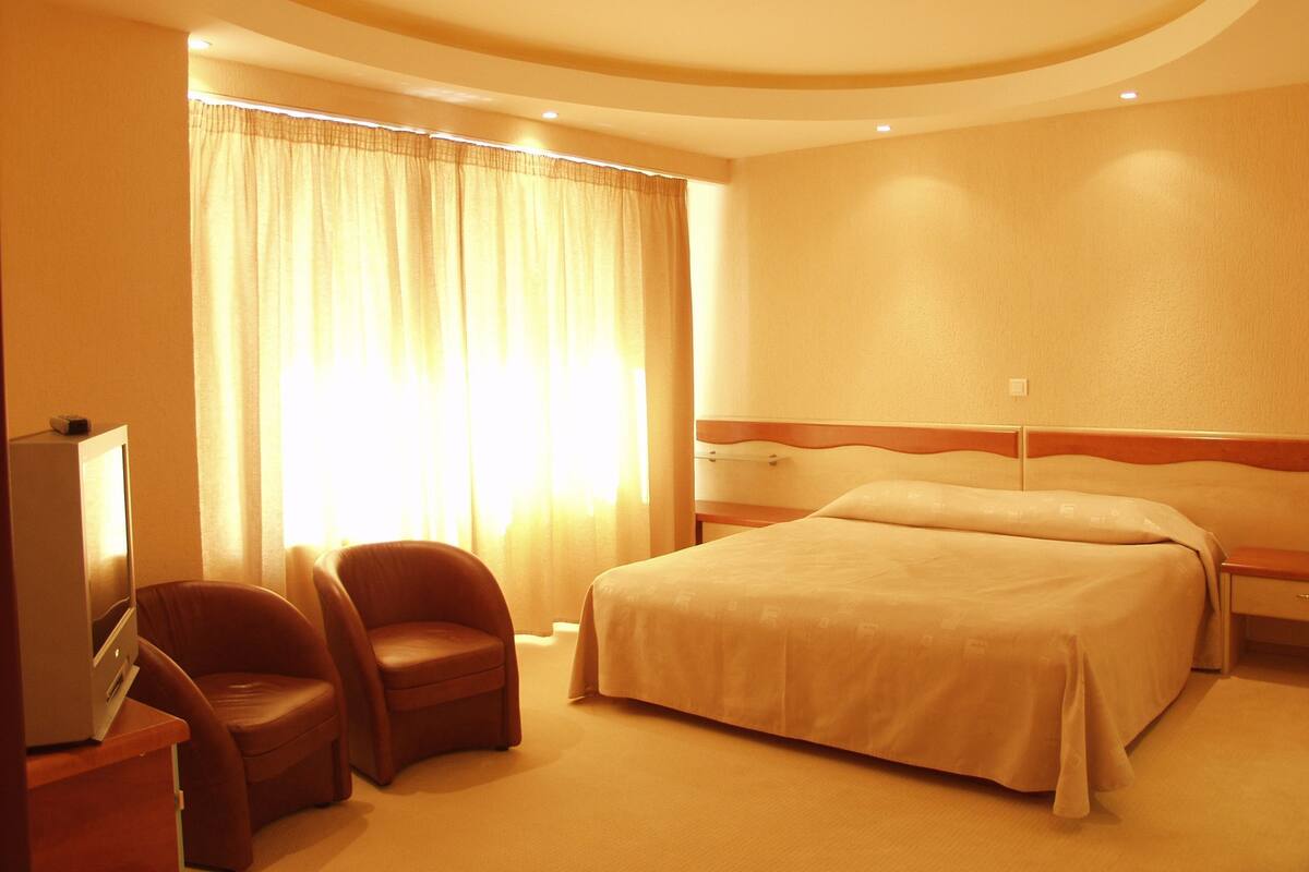 A hotel room with a bed, two armchairs, a television, and a large window with curtains.