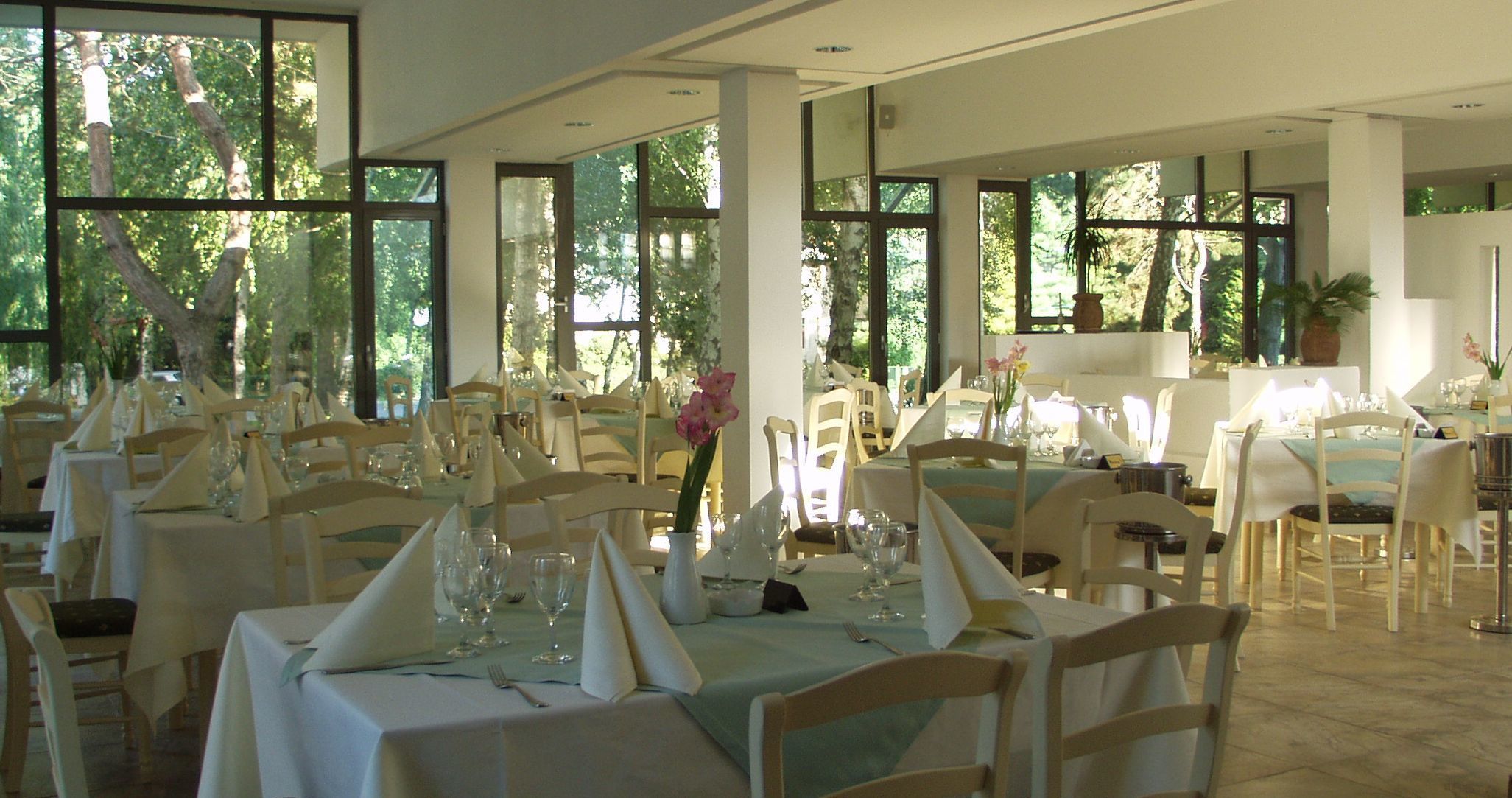 A restaurant with multiple tables set for dining, large windows with natural light, and a view of trees outside.