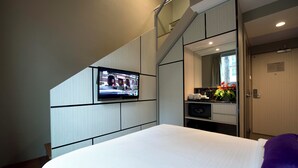 Ein Hotelzimmer mit Bett, Wandfernseher, einer kleinen Küchenzeile und einem Badezimmer mit Spiegel.