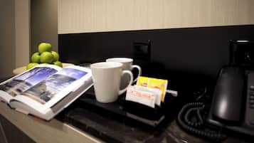 Un comptoir dans une chambre d’hôtel avec un magazine, une tasse blanche, un téléphone et un petit panier de fruits.