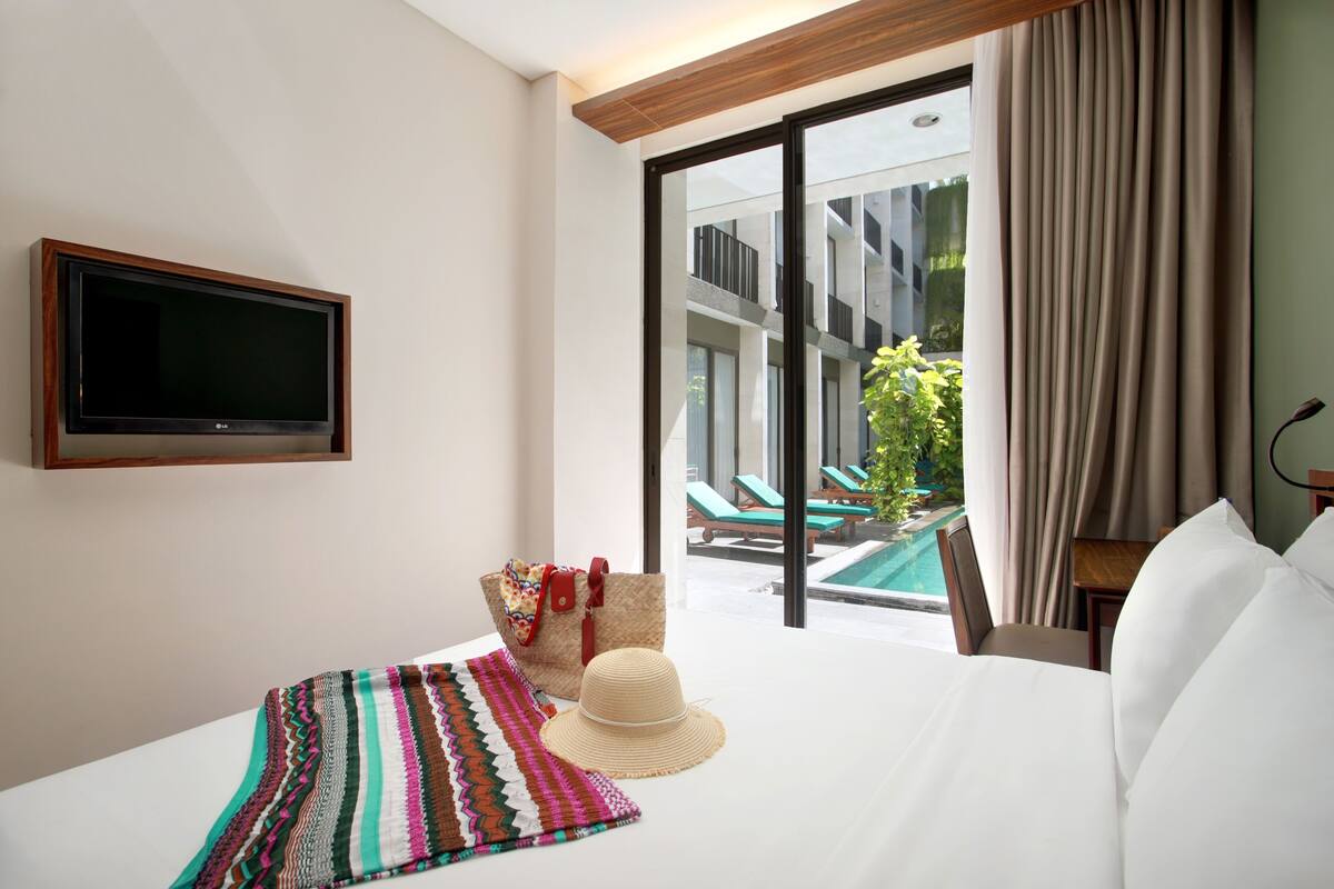 Une chambre d’hôtel avec un lit, une télévision, une vue sur une piscine et un espace extérieur aménagé pour s’asseoir, ainsi qu’une grande fenêtre.